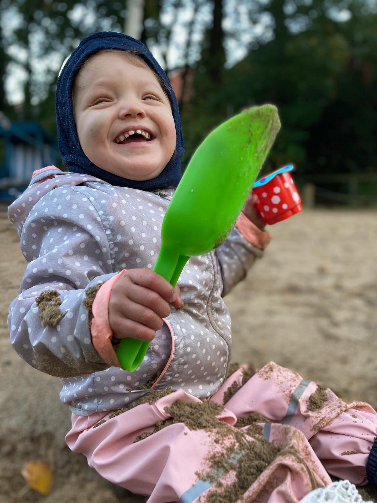 Line sitzt mit Matschhose, Jacke und Mütze und einer grünen Schaufel in der Hand auf einem Spielplatz im Sand und lacht