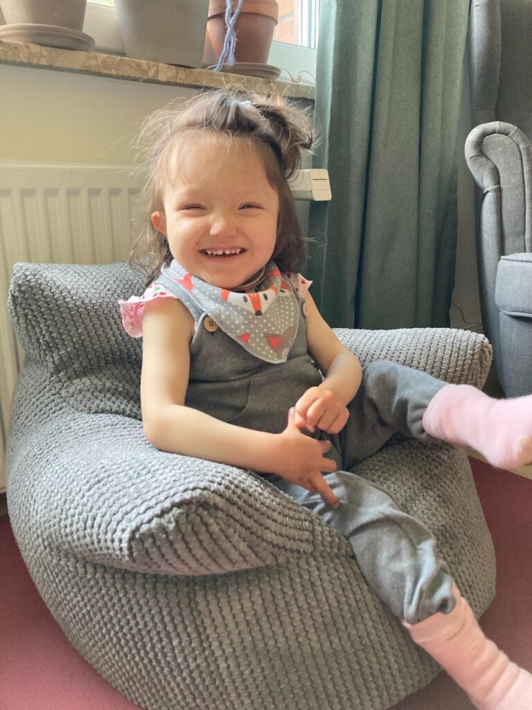 Line sitzt lachend auf einem grauen Kindersessel. Sie trägt eine graue Latzhose und rosa Socken und strahlt übers ganze Gesicht.
