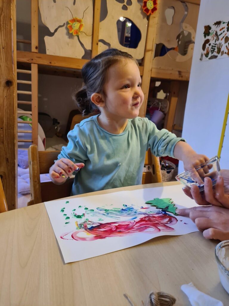 Line greift in eine Schale mit Fingermalfarbe, vor ihr liegt ein bereits teilweise bemaltes Blatt Papier.